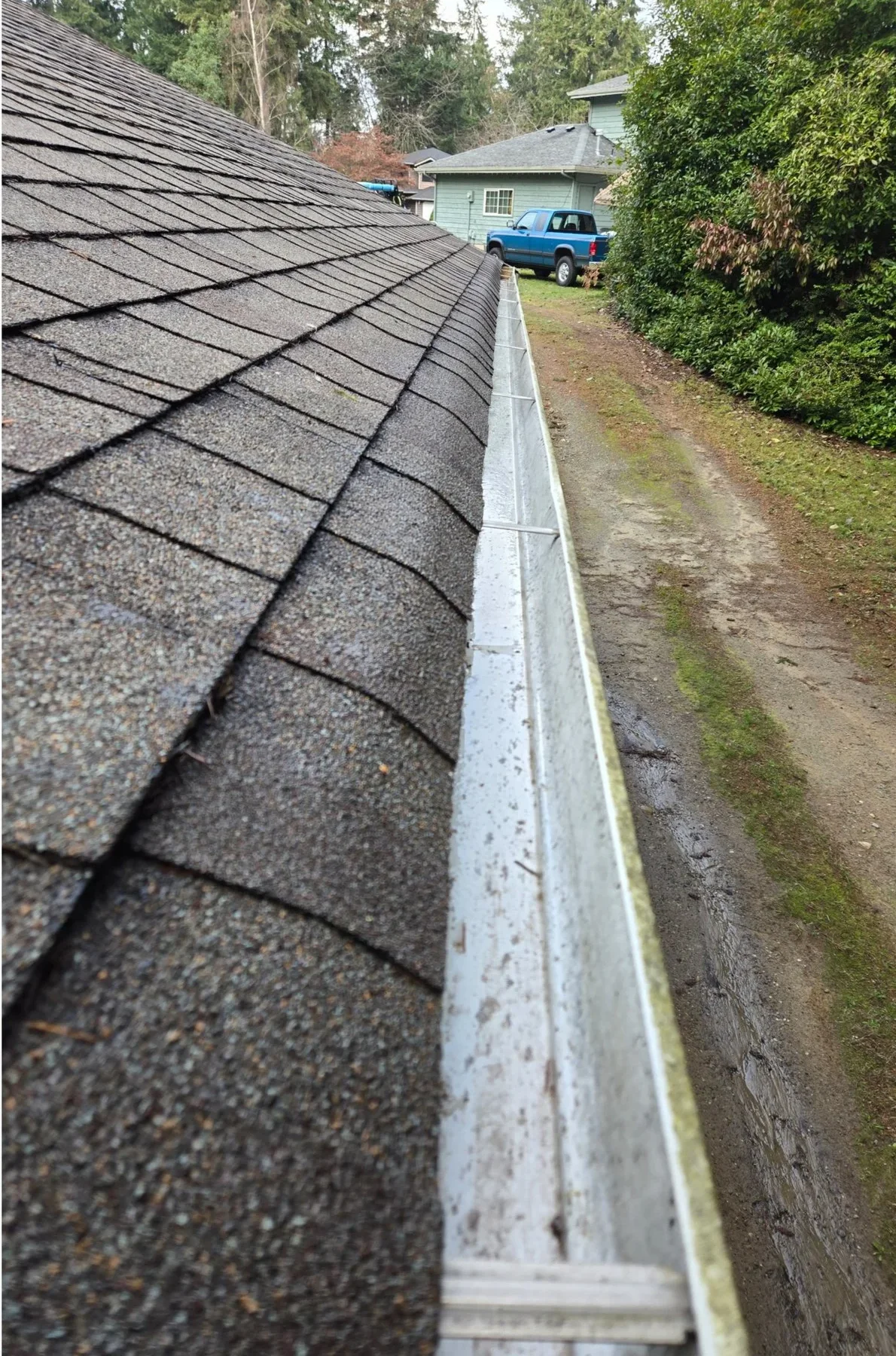 Close-up view of a shingled roof and metal gutter along the edge, with a blue pickup and house in the background.