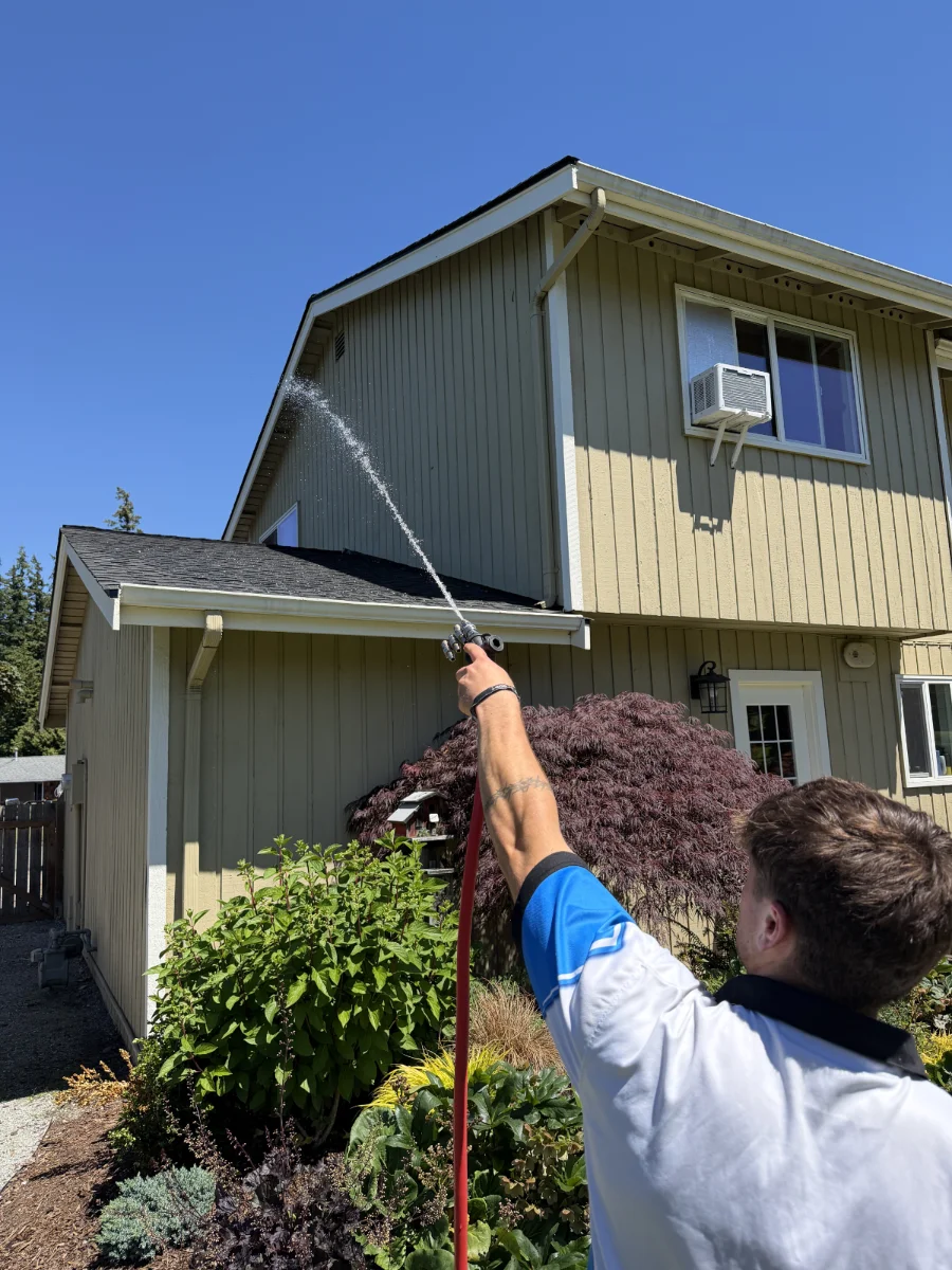 roof-cleaning-img-2 Person in a blue and white shirt pressure-washing the beige house siding under a clear blue sky.