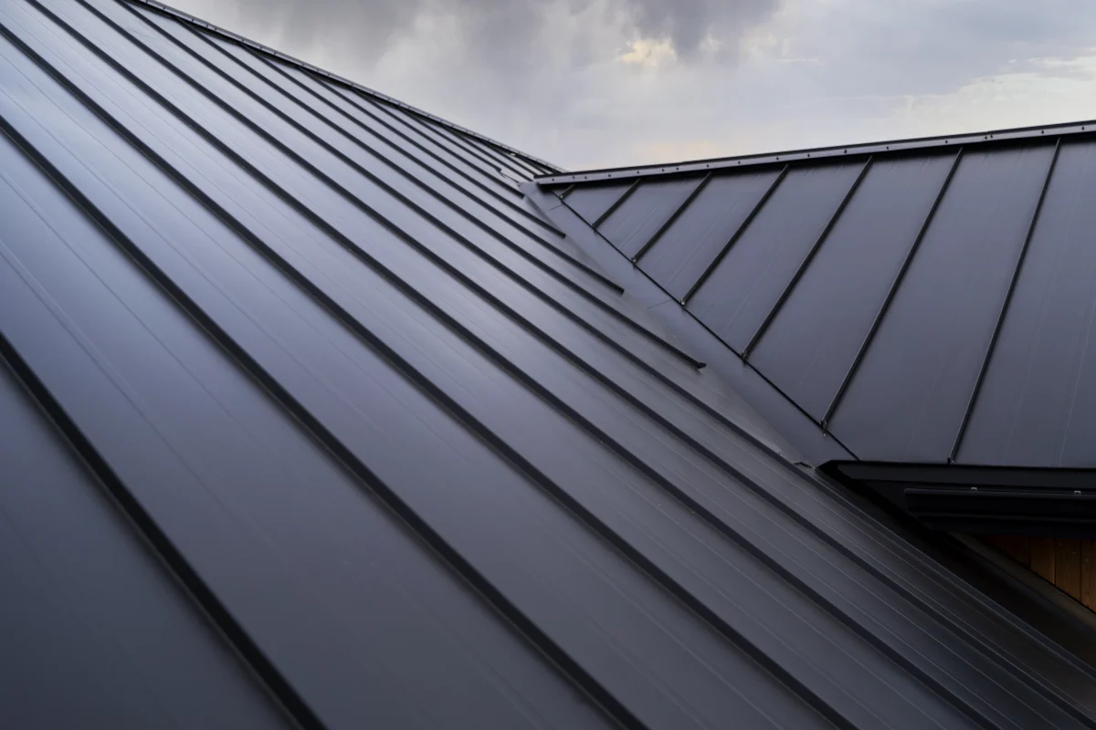 Close-up view of dark gray metal roofing panels meeting at a ridge under a cloudy sky.