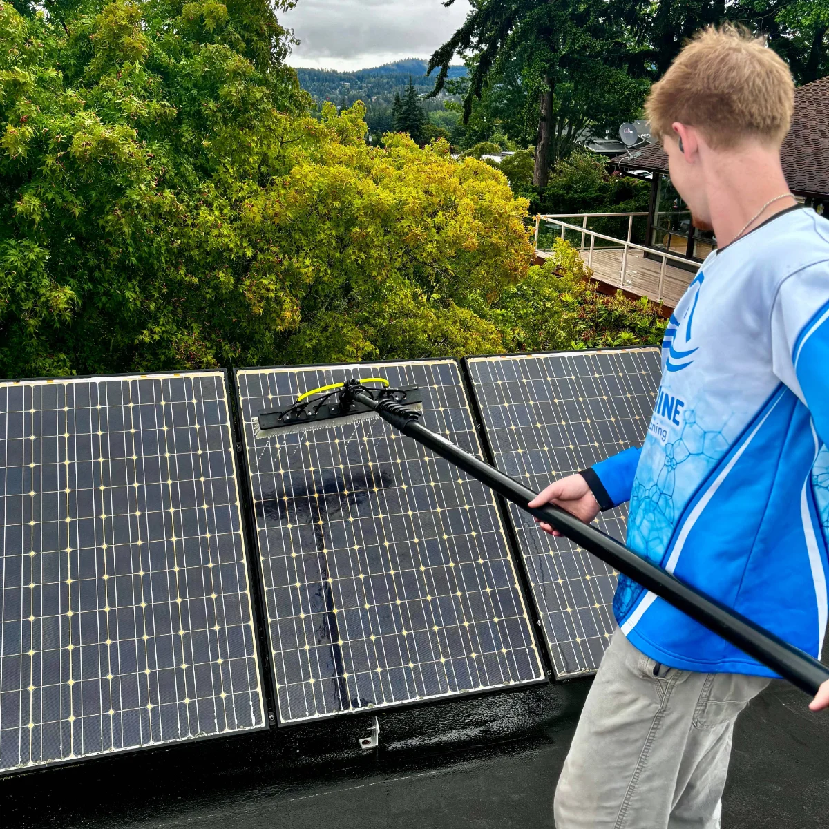 solar-panel-cleaning-img-1 Person cleaning solar panels on a rooftop with a long cleaning wand, amid green trees in the background.
