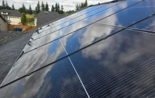 solar-panel-cleaning-img-3 Rows of glossy solar panels on a sloped roof, reflecting a blue sky and white clouds in a suburban neighborhood.