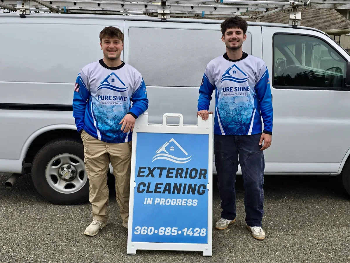 window-cleaning-img-4 Two workers in blue-and-white uniforms stand beside a van, posing with a sign that reads Exterior Cleaning In Progress and a phone number.